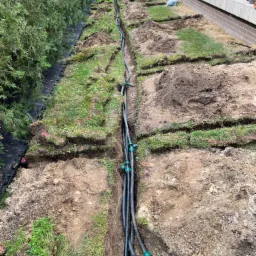 Wykopany rów w ziemi wzdłuż trawnika z częściowo ułożonymi czarnymi rurami z zielonymi złączkami, widoczny system nawadniania ogrodu w trakcie instalacji.