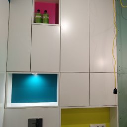 UmeblujSie - Biała zabudowa meblowa z kolorowymi wnękami (różowa, turkusowa, żółta) w łazience, widoczny fragment umywalki i instalacji elektrycznej.