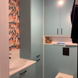 UmeblujSie - Niebieska zabudowa łazienkowa z szafkami, umywalką i toaletą, wzorzysta tapeta na ścianie, podłoga w stylu lastryko.