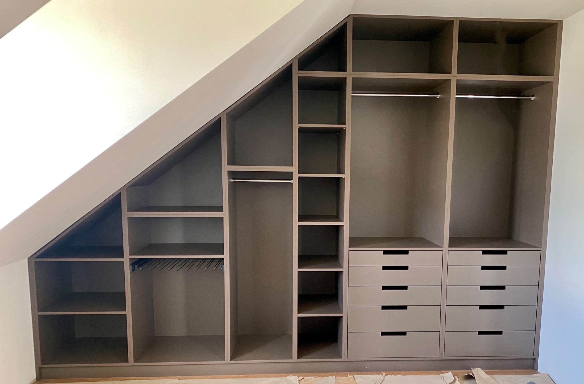 Szara szafa wnękowa na poddaszu ze skośnym sufitem, z półkami, drążkami na wieszaki i szufladami, widok na puste wnętrze.
