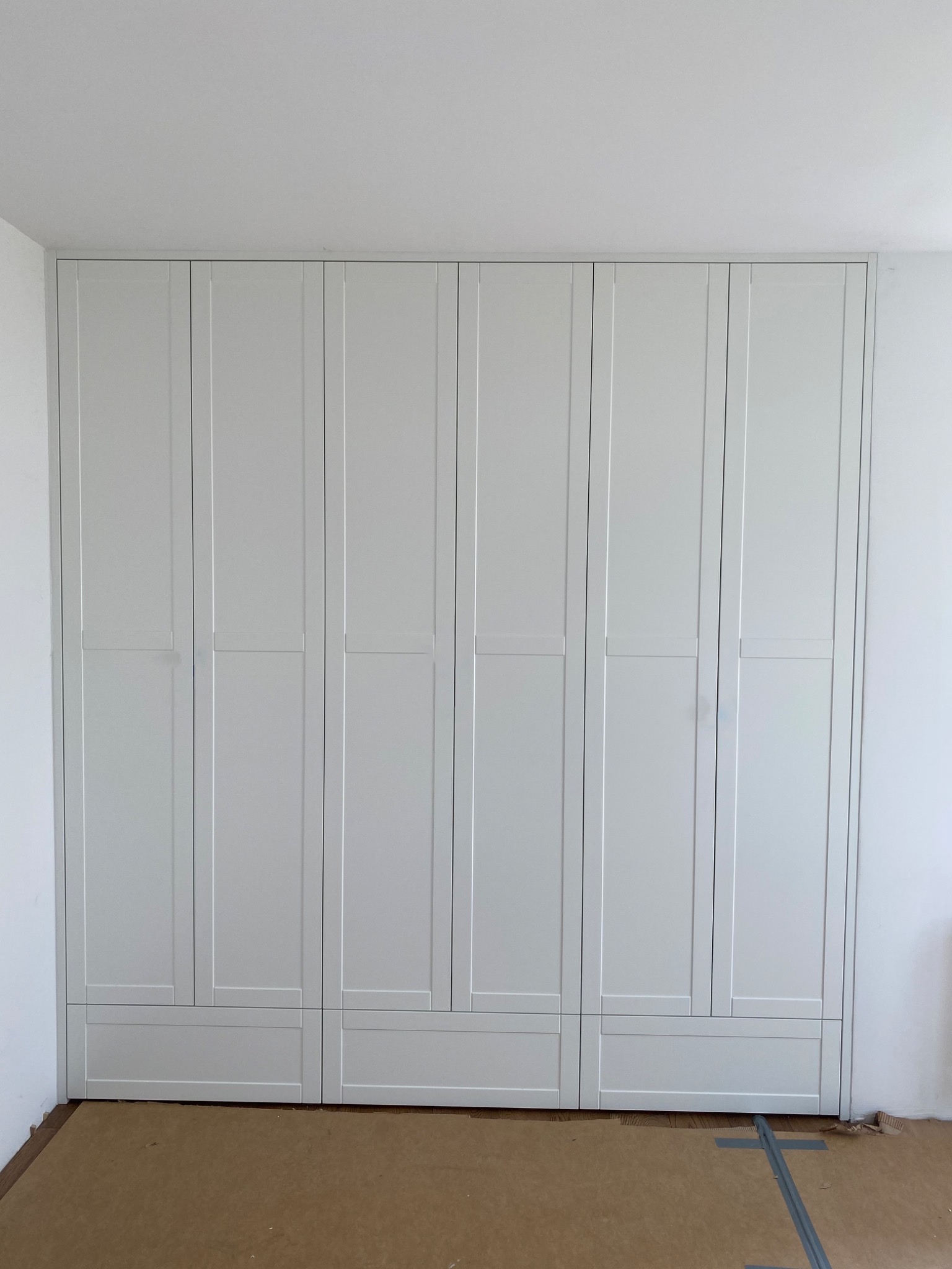 Biała szafa wnękowa z panelowymi drzwiami i szufladami u dołu, zabezpieczona kartonem podłoga w trakcie montażu.