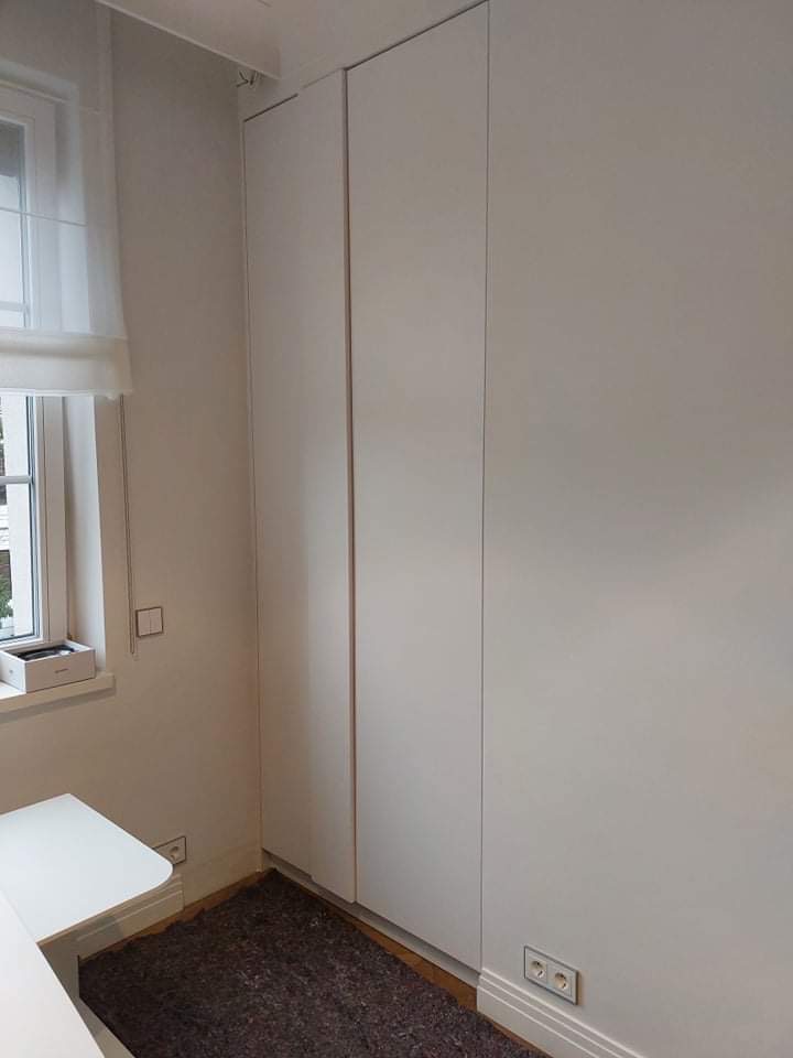 Biała, minimalistyczna szafa wnękowa z trzema drzwiami, dopasowana do koloru ścian, z widocznymi gniazdkami elektrycznymi na ścianie i dywanikiem na podłodze.