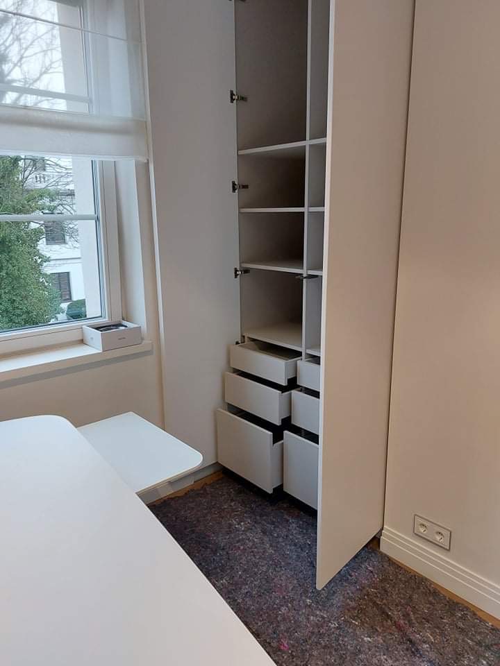 Wnętrze pokoju z białą, wysoką szafą wnękową z otwartymi drzwiami, widoczne półki i szuflady, obok okno z roletą rzymską i biały stolik.