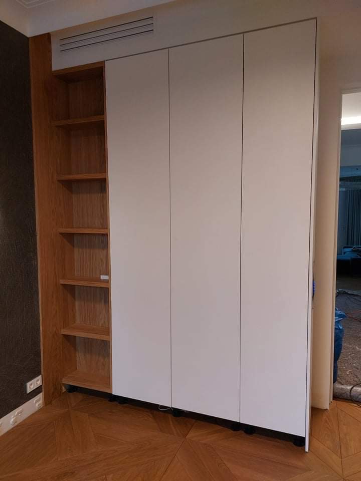 Wbudowana szafa z białymi frontami i otwartym regałem z jasnego drewna, widoczna na tle parkietu w jodełkę.