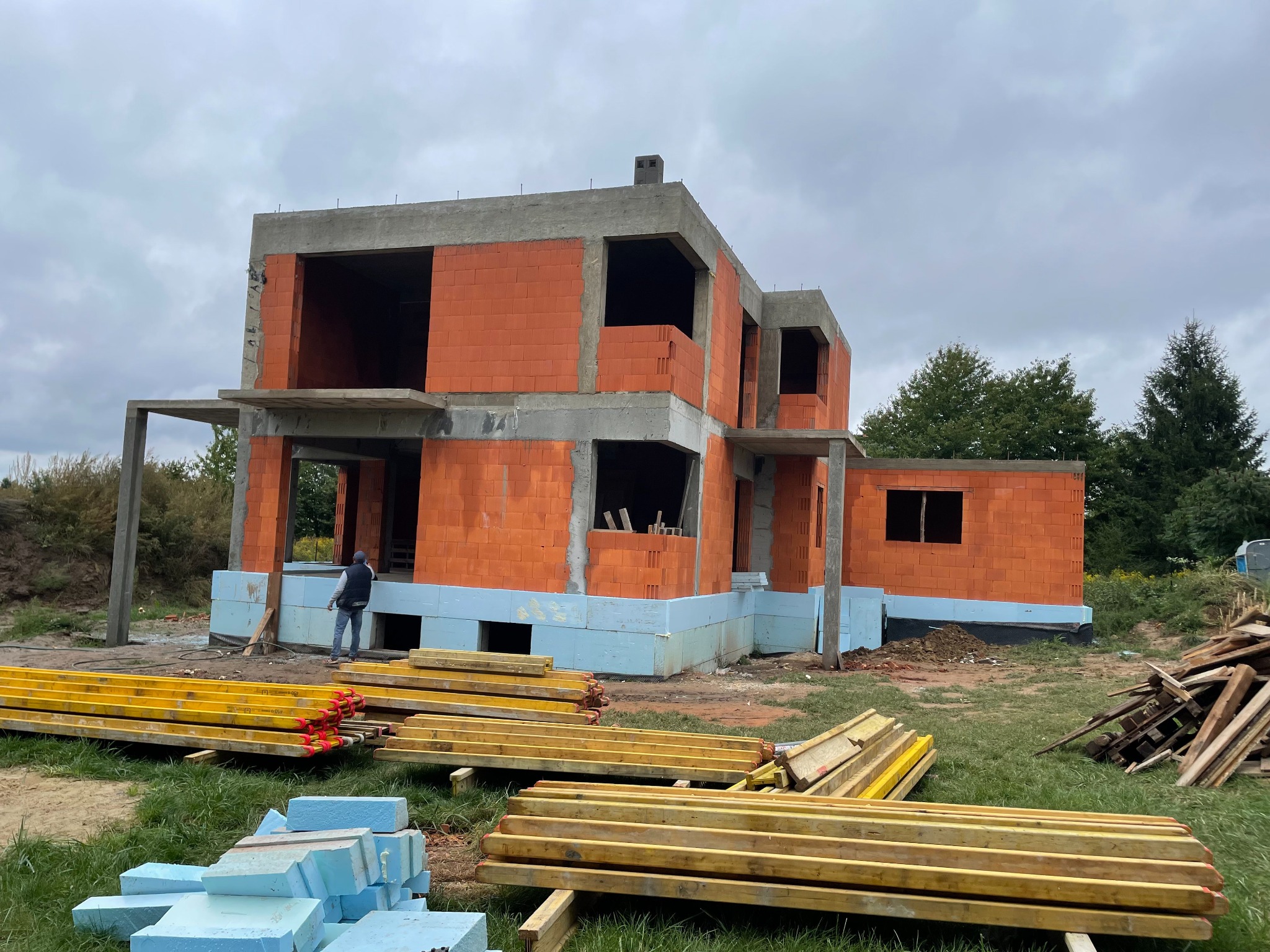 Dwukondygnacyjny dom w trakcie budowy z czerwonej cegły, z widocznymi elementami szalunkowymi i izolacją fundamentów, na pierwszym planie składowane materiały budowlane i pracownik w oddali.