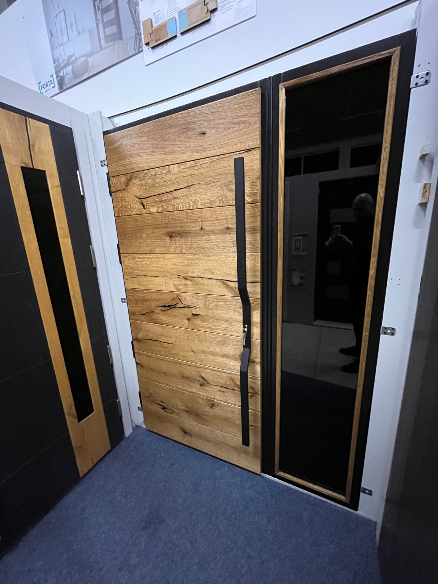 Nowoczesne drzwi zewnętrzne z drewna o naturalnym usłojeniu, z czarnym, pionowym uchwytem i czarną ramą, obok drzwi z czarnymi panelami i jasnymi wstawkami, na podłodze niebieska wykładzina, w tle...
