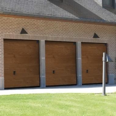 Trzy brązowe, segmentowe bramy garażowe z fakturą drewna, umieszczone obok siebie w ceglanym budynku, widoczne z perspektywy trawnika.