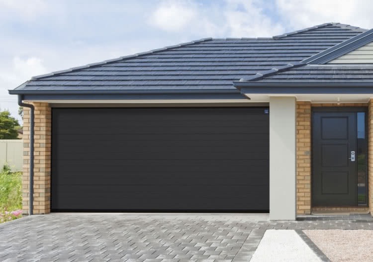 Nowoczesna, grafitowa brama garażowa segmentowa z poziomymi przetłoczeniami, zamontowana w budynku z ceglaną elewacją i szarą dachówką.