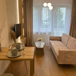 Jasne, nowocześnie urządzone wnętrze salonu z drewnianą podłogą, beżową sofą i stołem z nakryciem na dwie osoby, oświetlone kulistymi lampami wiszącymi i ledowym oświetleniem ściennym.