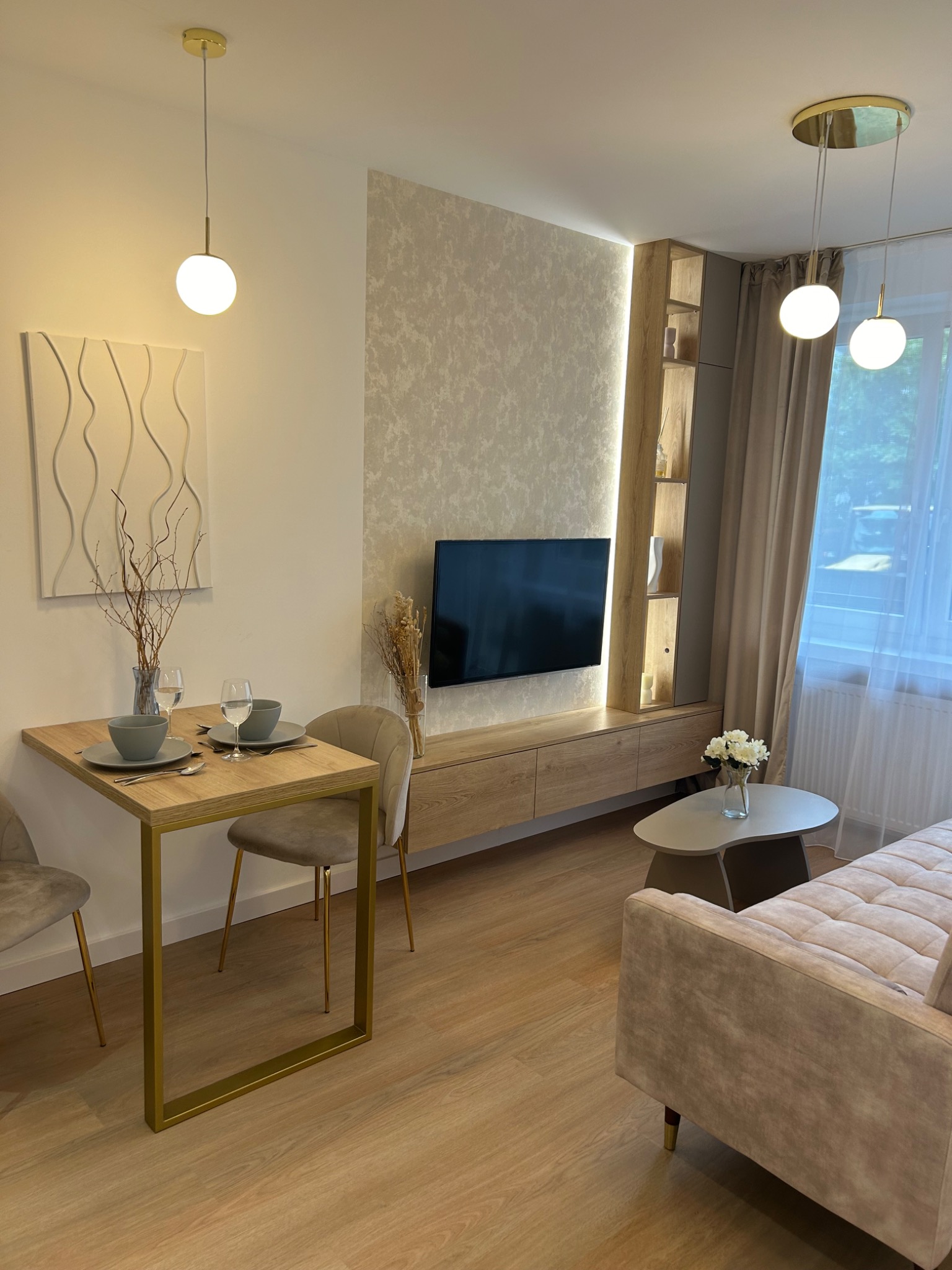 Nowoczesne wnętrze salonu z drewnianą podłogą, ścianą akcentową z tapetą, telewizorem, szafką RTV, stolikiem kawowym z kwiatami i stołem jadalnym z zastawą, oświetlone kulistymi lampami wiszącymi.
