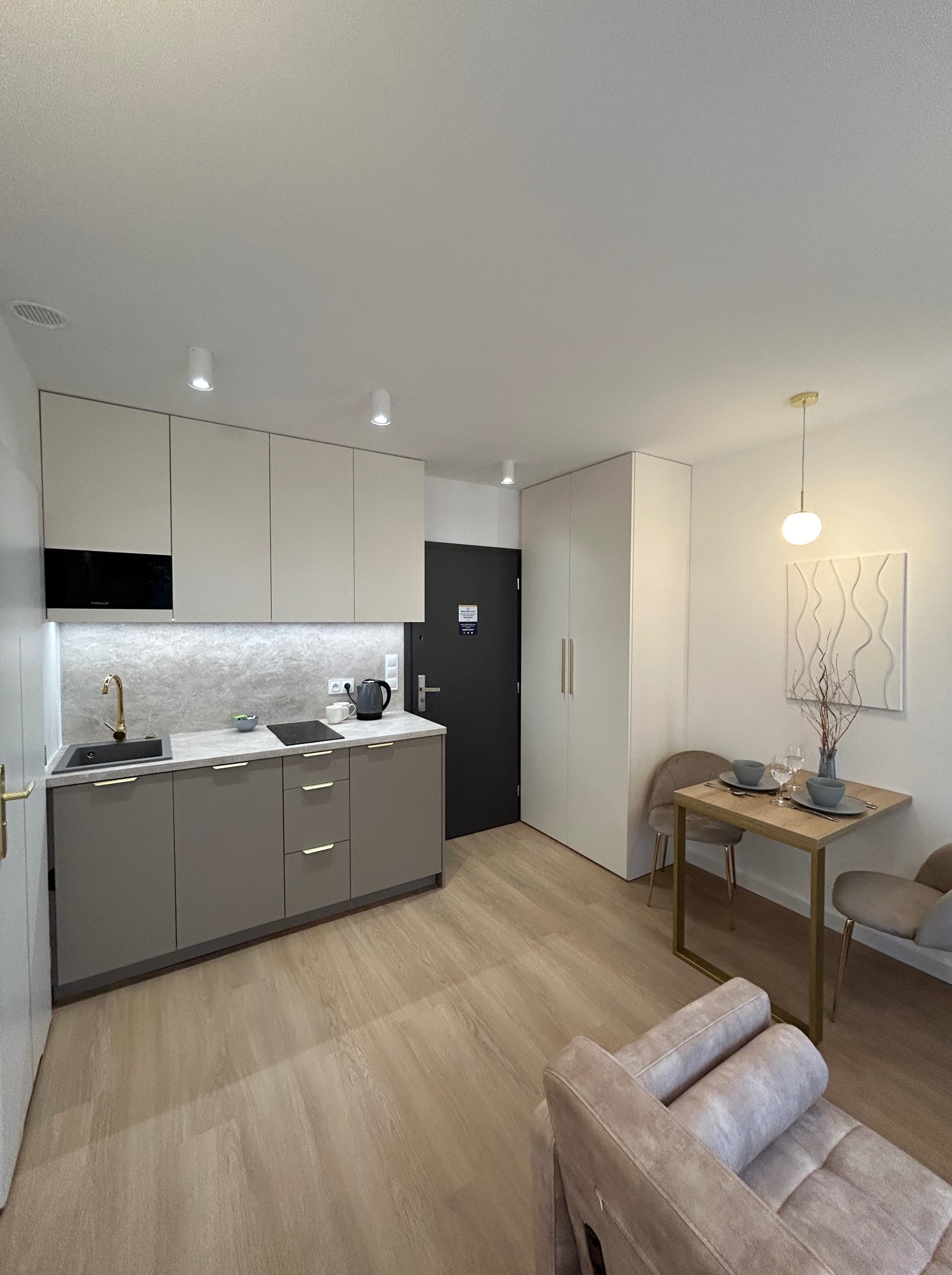 Wnętrze nowoczesnej kawalerki z aneksem kuchennym w odcieniach beżu i szarości, drewniana podłoga, stół z zastawą dla dwóch osób i minimalistyczna dekoracja ścienna.