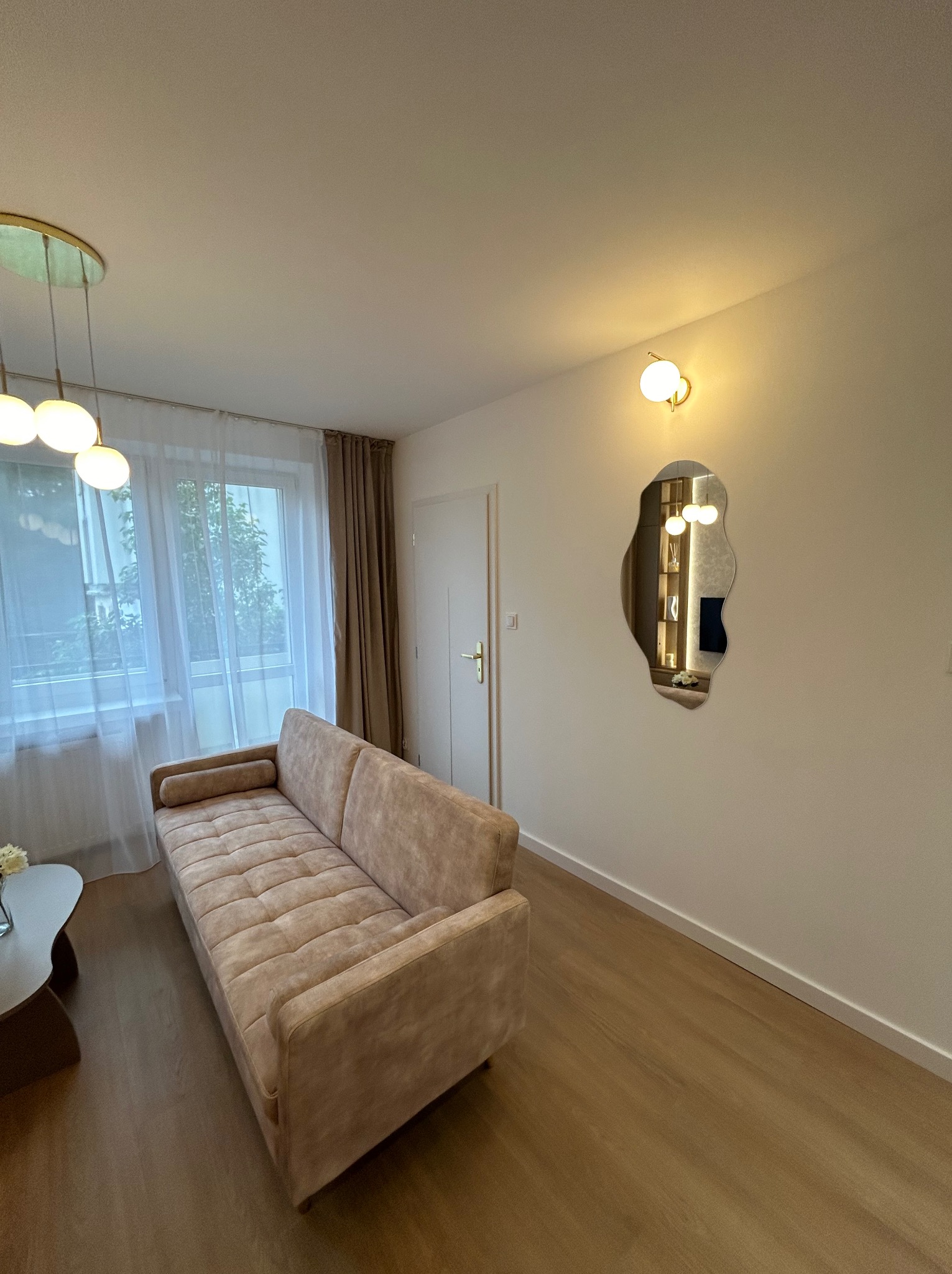 Jasny salon z beżową sofą pod oknem z białymi zasłonami, parkietem i asymetrycznym lustrem na ścianie odbijającym oświetloną półkę z dekoracjami.