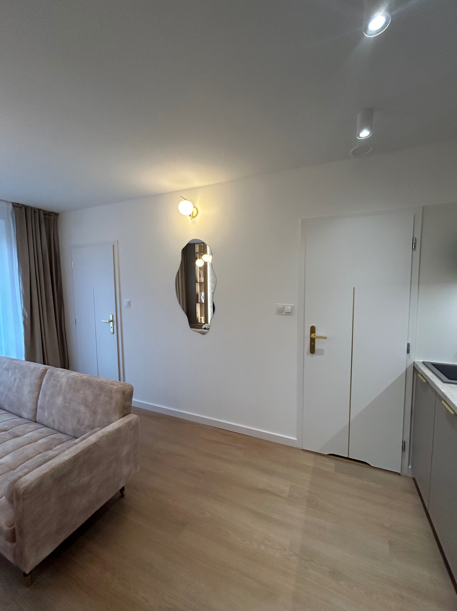 Jasne wnętrze salonu z beżową sofą, drewnianą podłogą, białymi drzwiami z minimalistycznymi złotymi klamkami i nieregularnym lustrem na ścianie obok lampy.