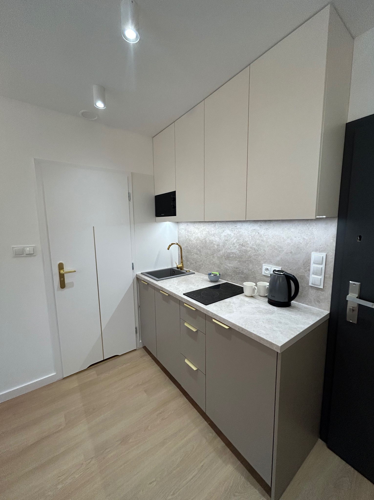 Nowoczesna, minimalistyczna kuchnia w odcieniach szarości i bieli, z złotymi akcentami na baterii i uchwytach szafek, oświetlona punktowymi lampami sufitowymi, z czajnikiem elektrycznym i dwoma...