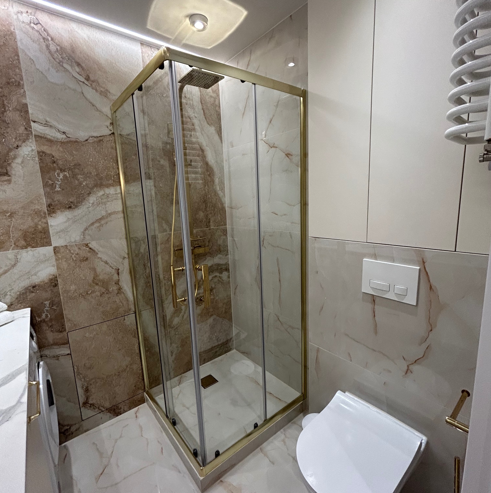 Kabina prysznicowa z złotymi profilami w nowoczesnej łazience z marmurowymi płytkami i grzejnikiem drabinkowym.