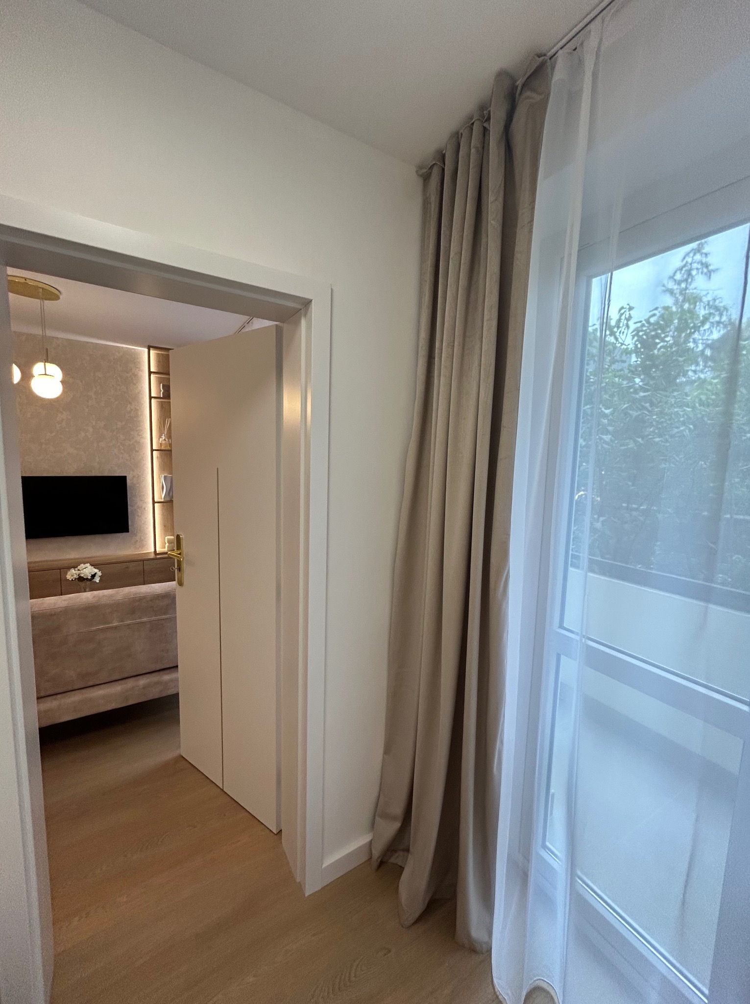 Ujęcie z korytarza na salon z beżowymi zasłonami i firankami w oknie, widoczny telewizor, szafka RTV i sofa w jasnych barwach.