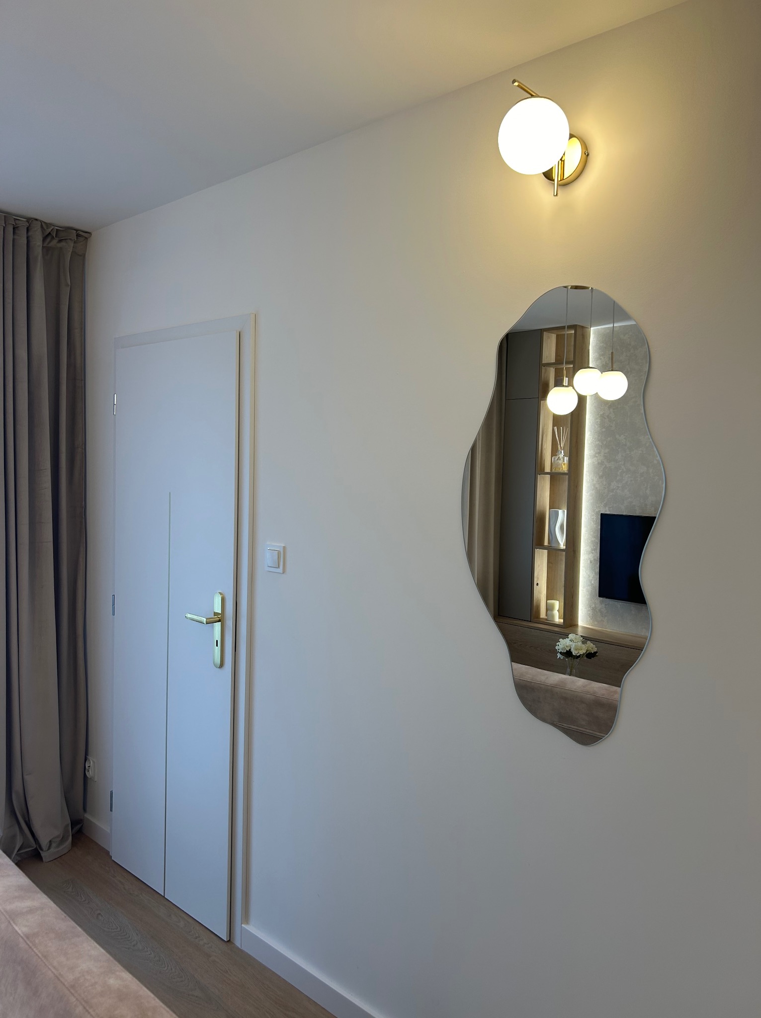 Nowoczesne wnętrze: białe drzwi z złotą klamką, designerskie lustro o nieregularnym kształcie odbijające oświetlony regał z dekoracjami, oraz kinkiet z białą kulą, na tle beżowej ściany.