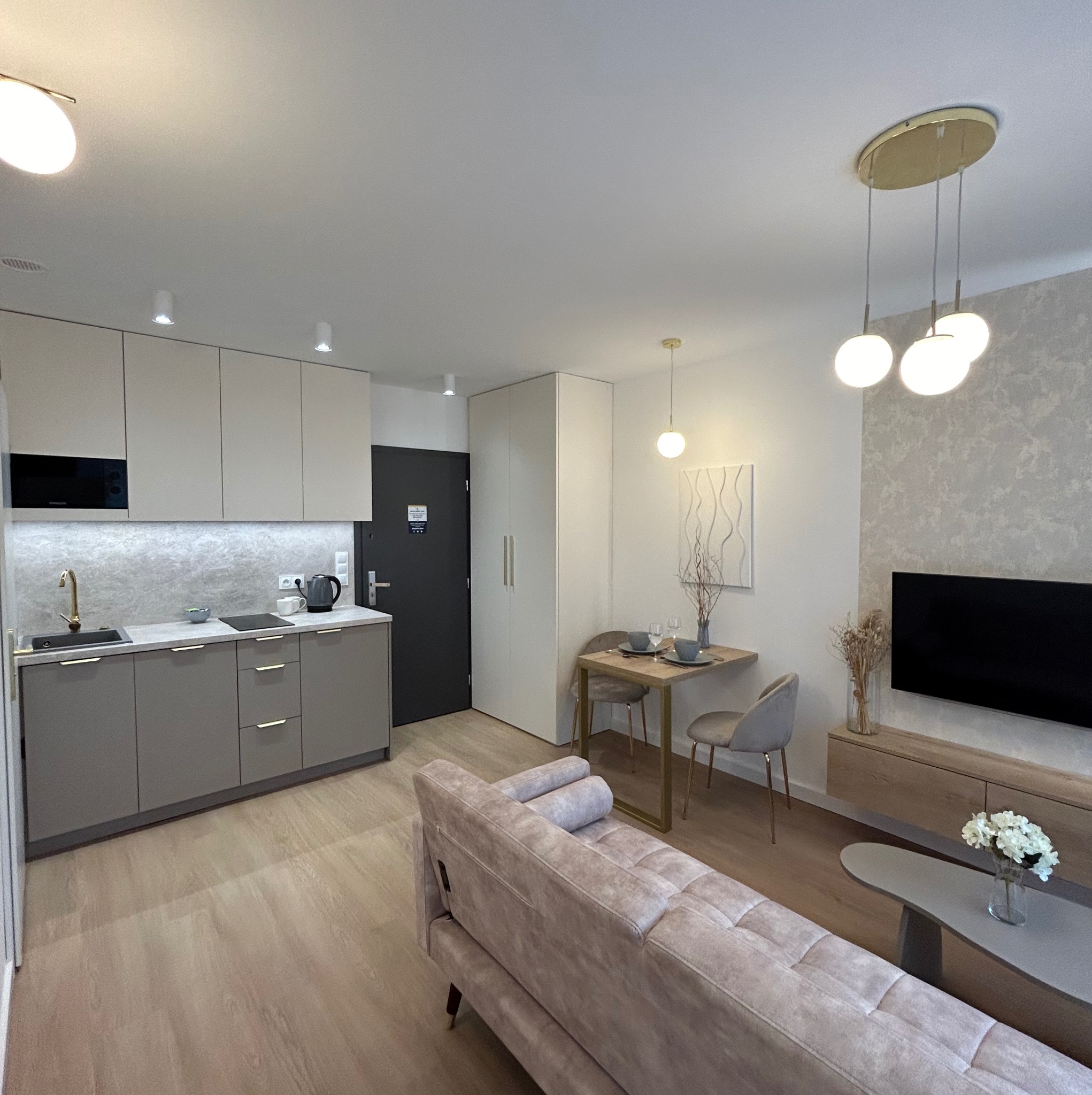 Nowoczesne wnętrze mieszkania z aneksem kuchennym, stołem jadalnym i sofą w odcieniach szarości i beżu, oświetlone lampami wiszącymi i sufitowymi.