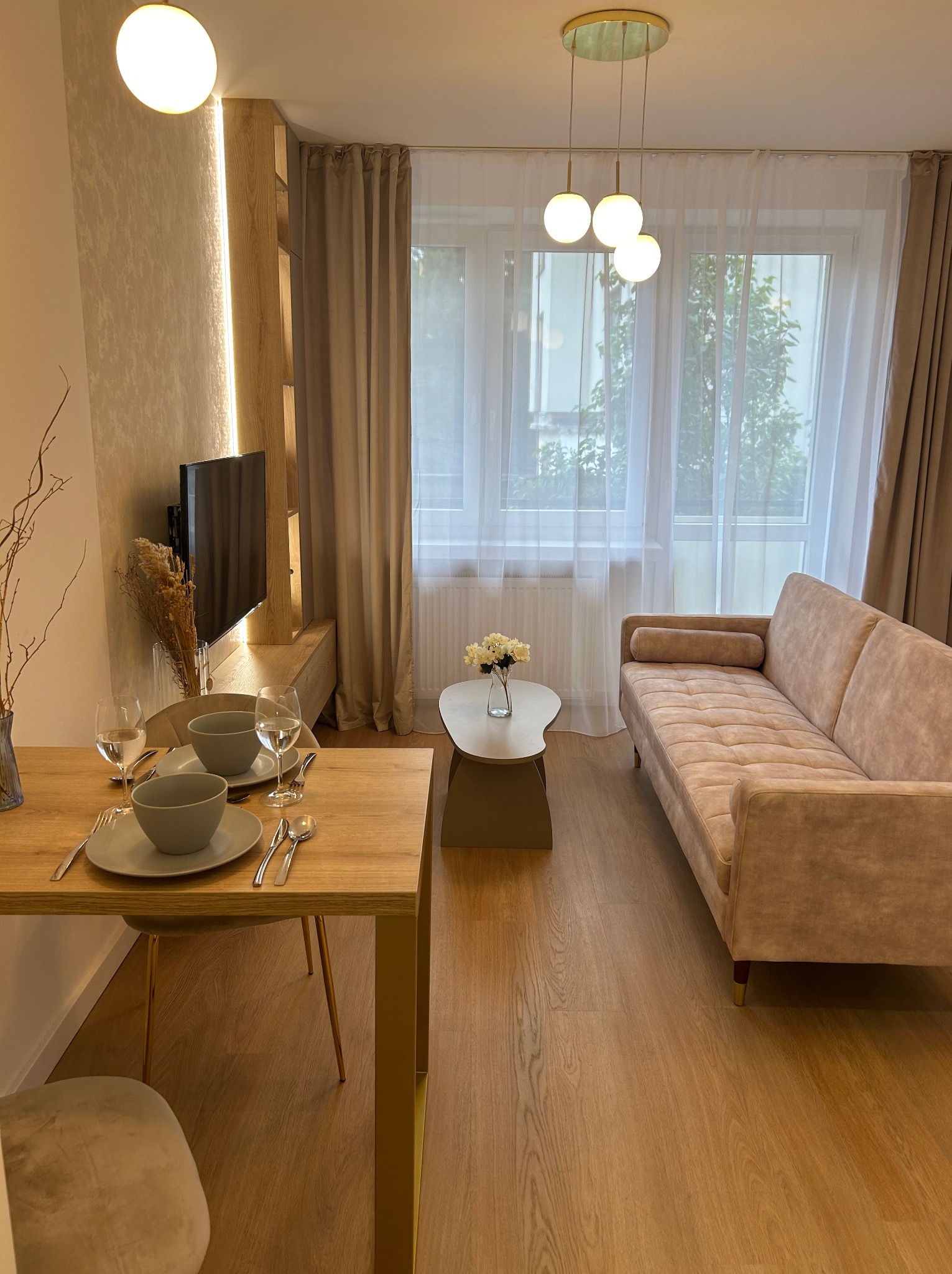 Jasne, nowocześnie urządzone wnętrze salonu z drewnianą podłogą, beżową sofą i stołem z nakryciem na dwie osoby, oświetlone kulistymi lampami wiszącymi i ledowym oświetleniem ściennym.