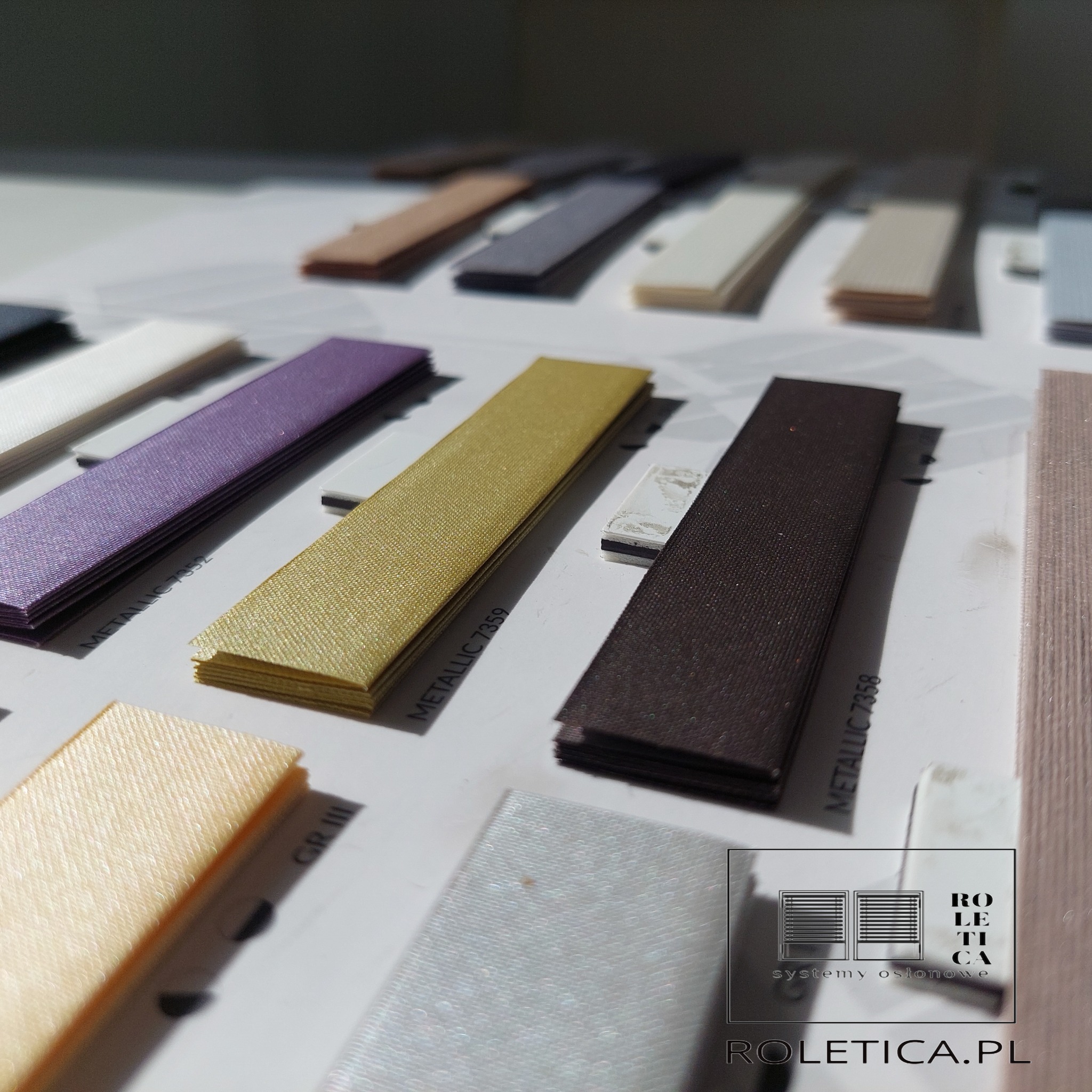 Próbki materiałów plisowanych w różnych kolorach, ułożone na białym tle, z widocznym logo firmy Roletica.pl w prawym dolnym rogu.