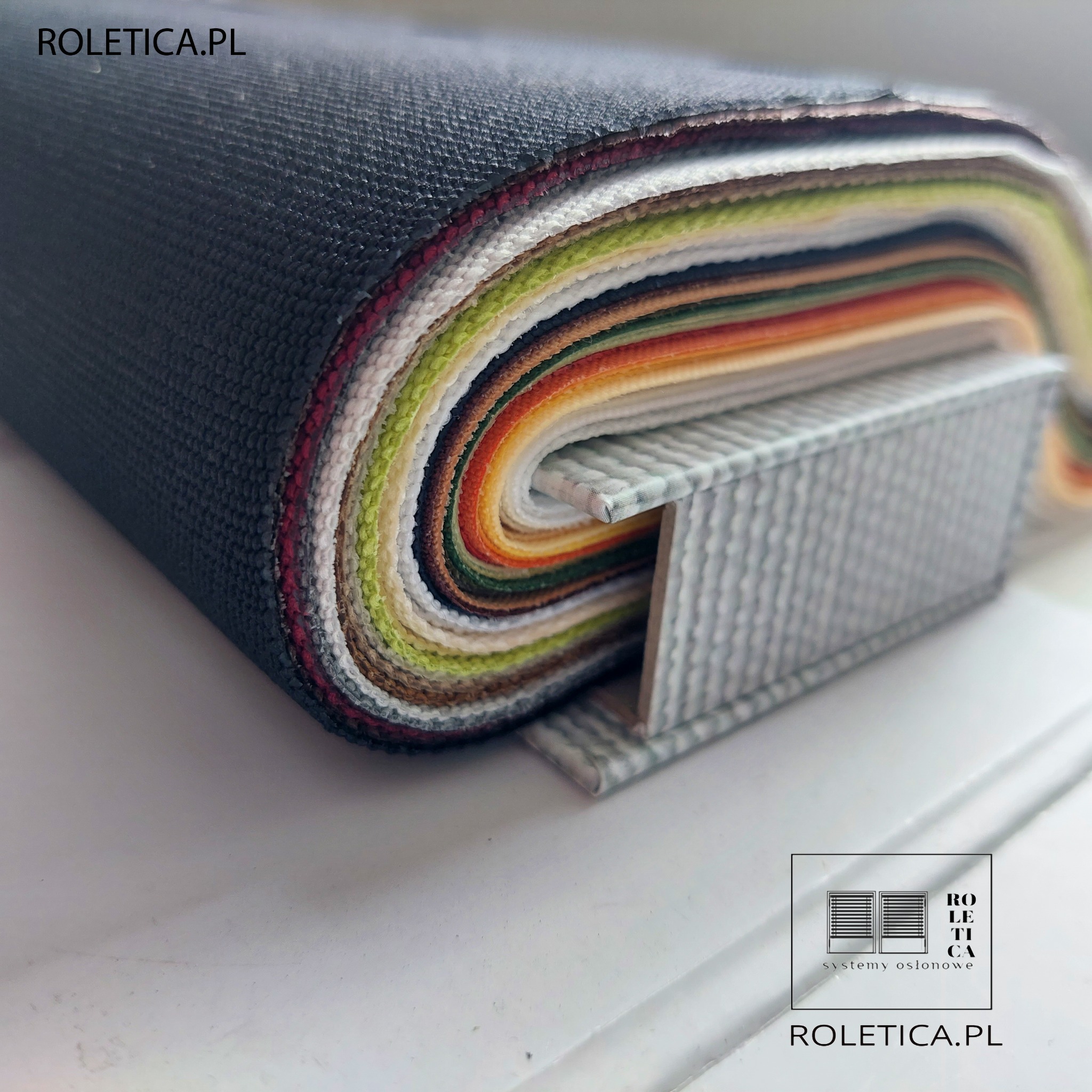 Przekrój przez zwinięte próbki tkanin w różnych kolorach i fakturach, używanych do produkcji systemów osłonowych, z widocznym logo firmy Roletica.pl.