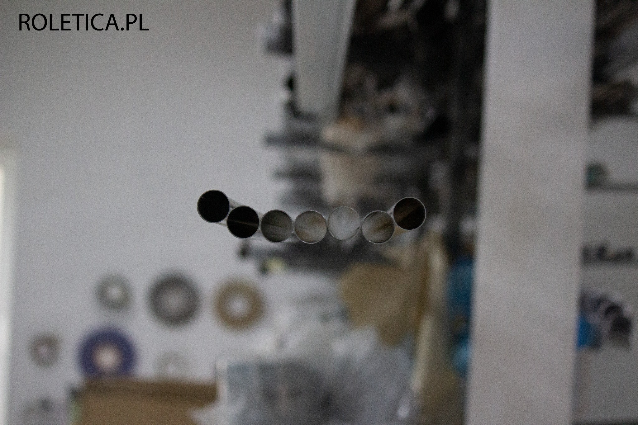Rury nawojowe do rolet, widoczne przekroje poprzeczne, w tle elementy warsztatu i logo Roletica.pl