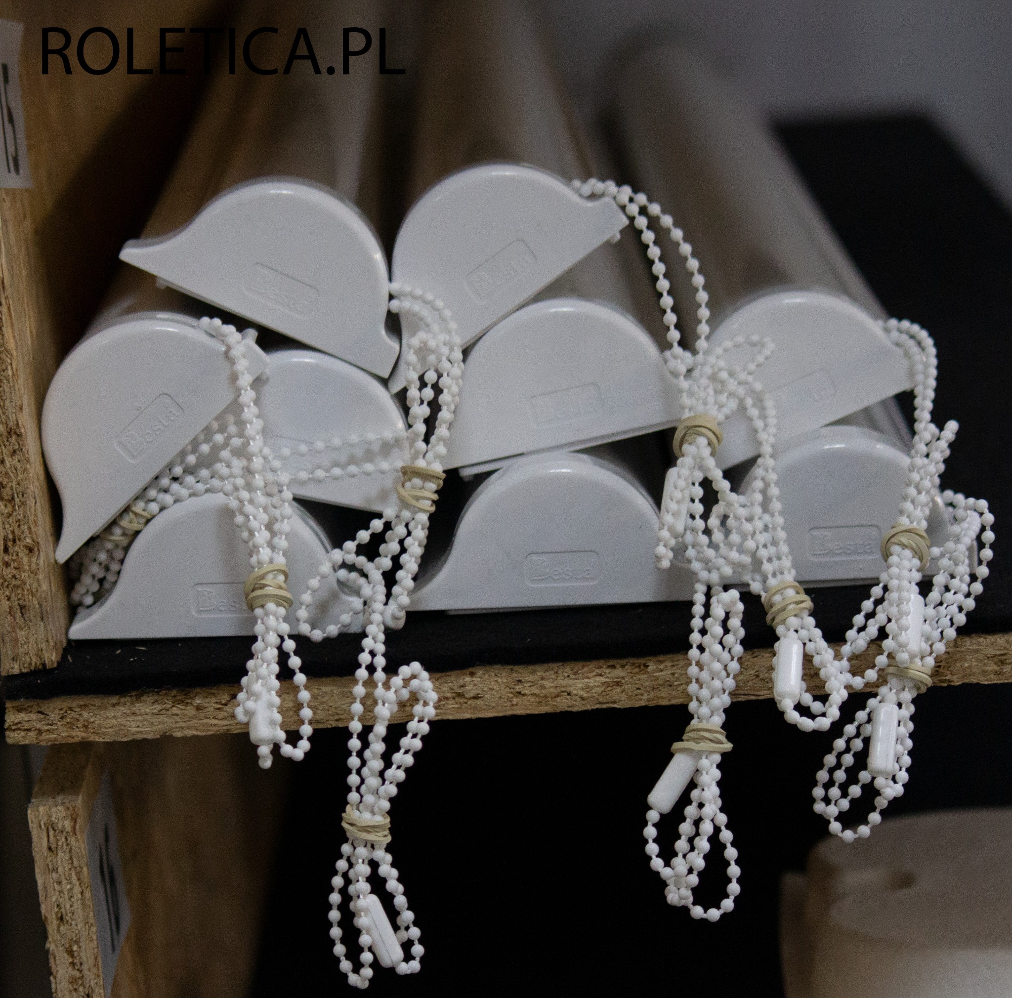 Stos kaset do rolet wewnętrznych marki Besta z łańcuszkami, zabezpieczonych gumkami recepturkami, ułożonych na półce w magazynie.