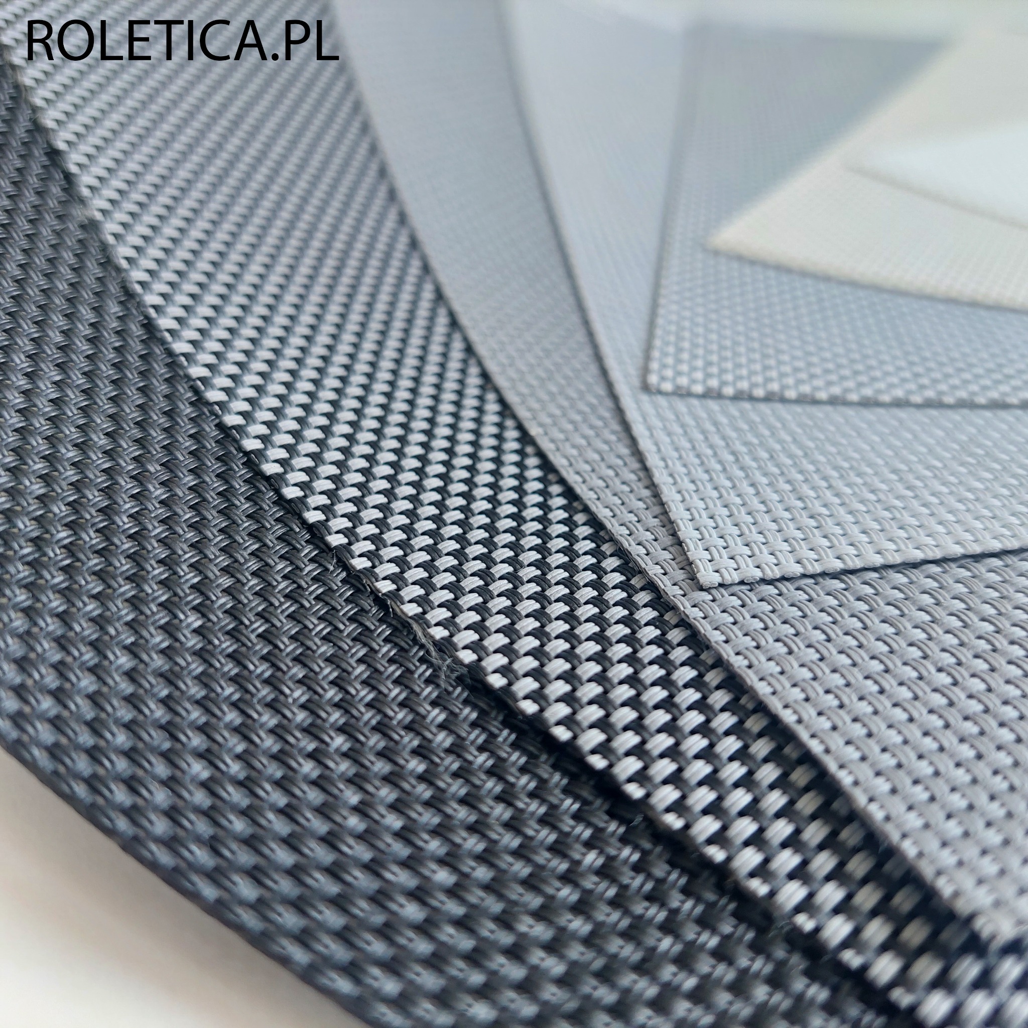 Zbliżenie na próbki tkanin o różnej fakturze i odcieniach szarości, prawdopodobnie do zastosowania w systemach zaciemniających okna, z widocznym logo Roletica.pl w lewym górnym rogu.