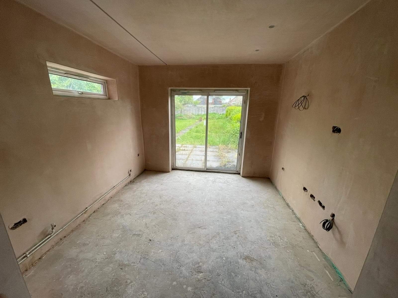 Pokój w trakcie remontu: ściany po nałożeniu gładzi, widoczne kable elektryczne, okno i przesuwne drzwi balkonowe z widokiem na ogród.