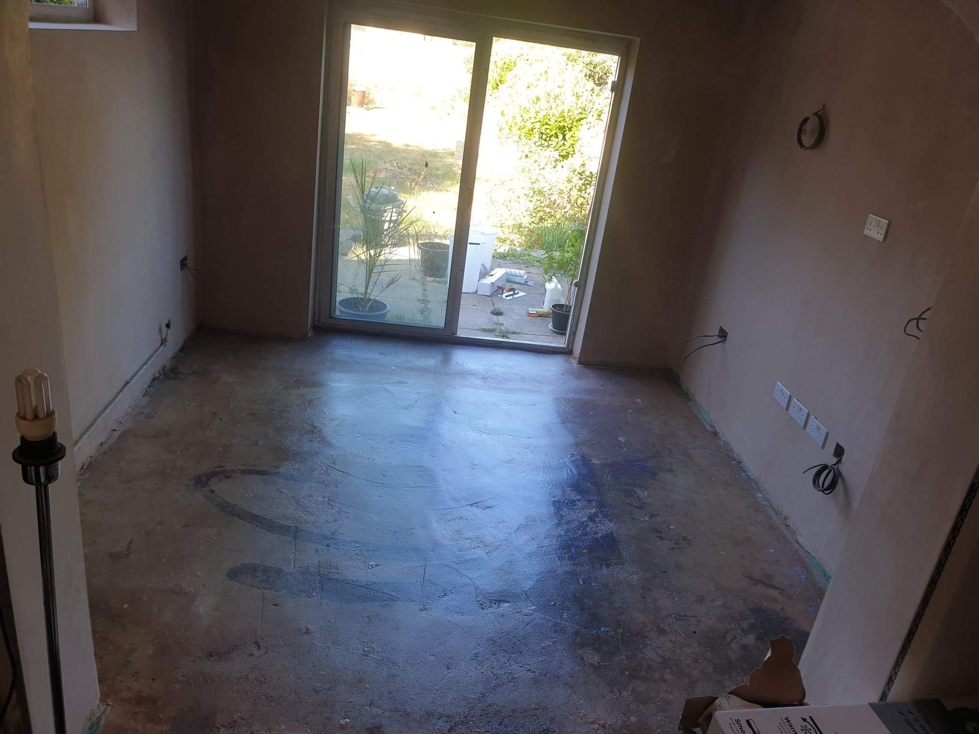 Pokój w trakcie remontu: surowe ściany po tynkowaniu, przygotowywane podłoże pod posadzkę, widoczne gniazdka elektryczne bez osprzętu, na zewnątrz przez przesuwne drzwi taras.