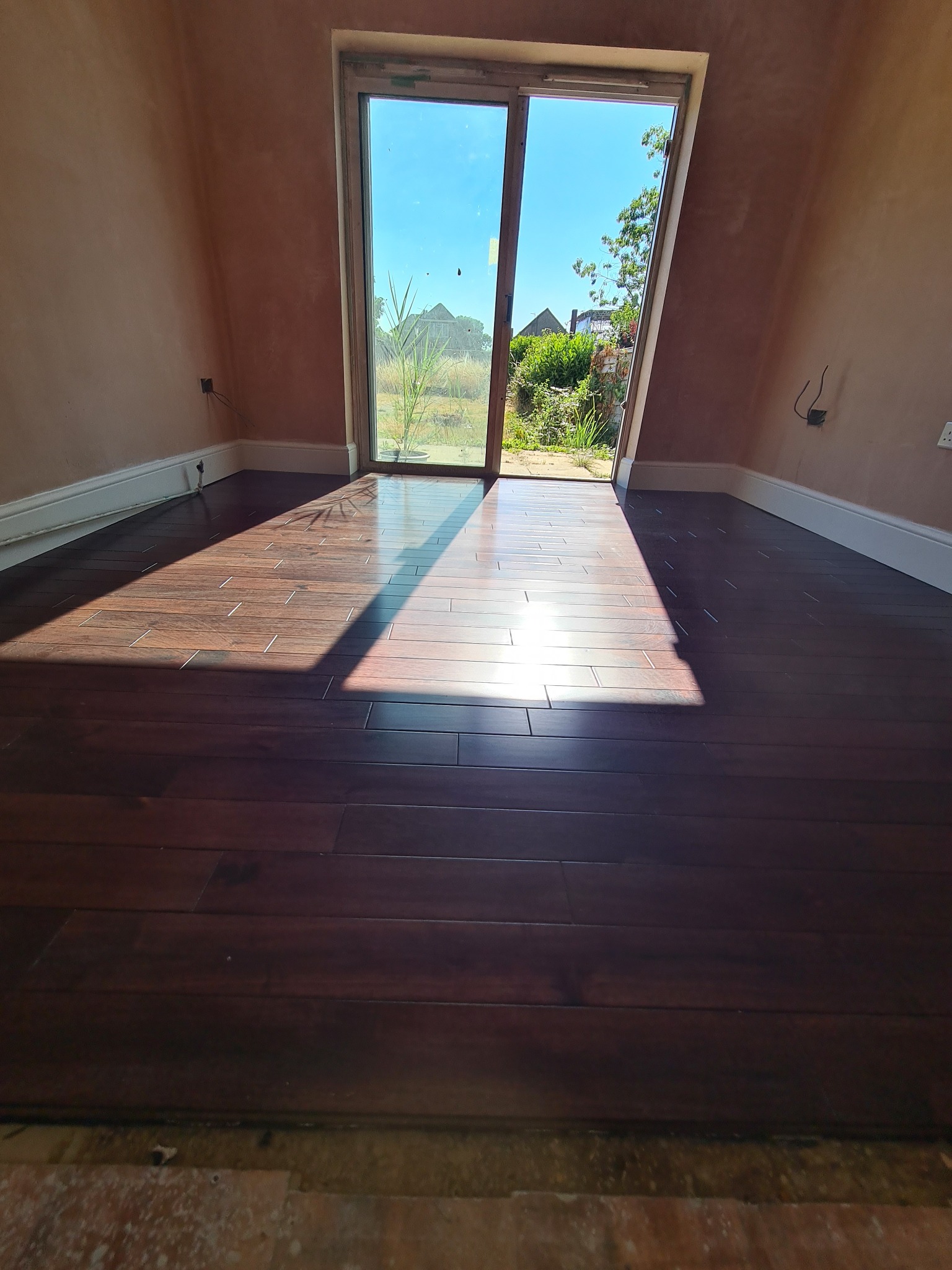 Ciemnobrązowa podłoga z paneli ułożona w pokoju z surowymi ścianami, widok na ogród przez przesuwne drzwi balkonowe, silne promienie słoneczne padają na podłogę.