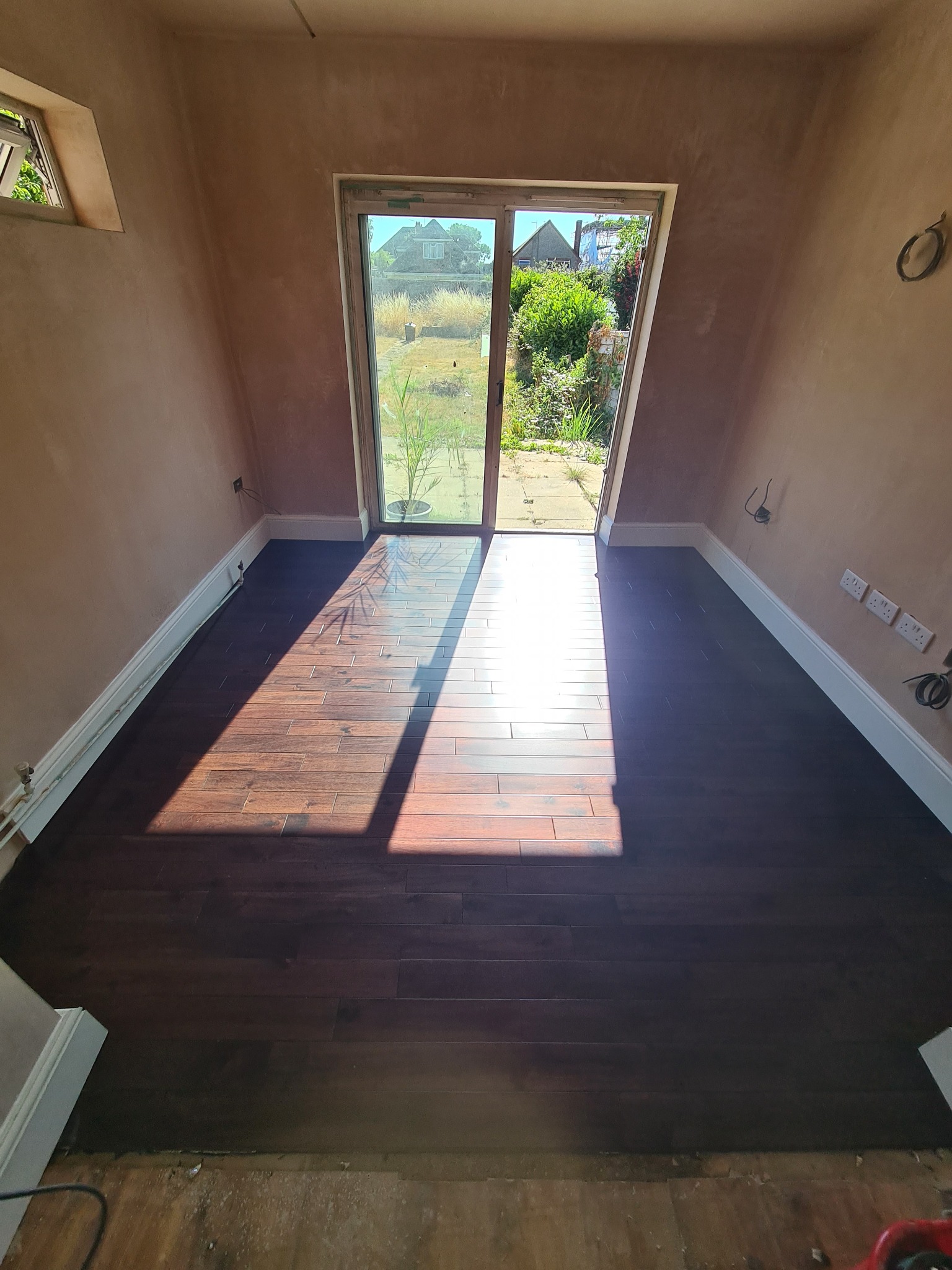 Nowo ułożona, ciemna podłoga z desek w pokoju z surowymi ścianami i dużym oknem balkonowym, przez które wpada silne światło słoneczne.