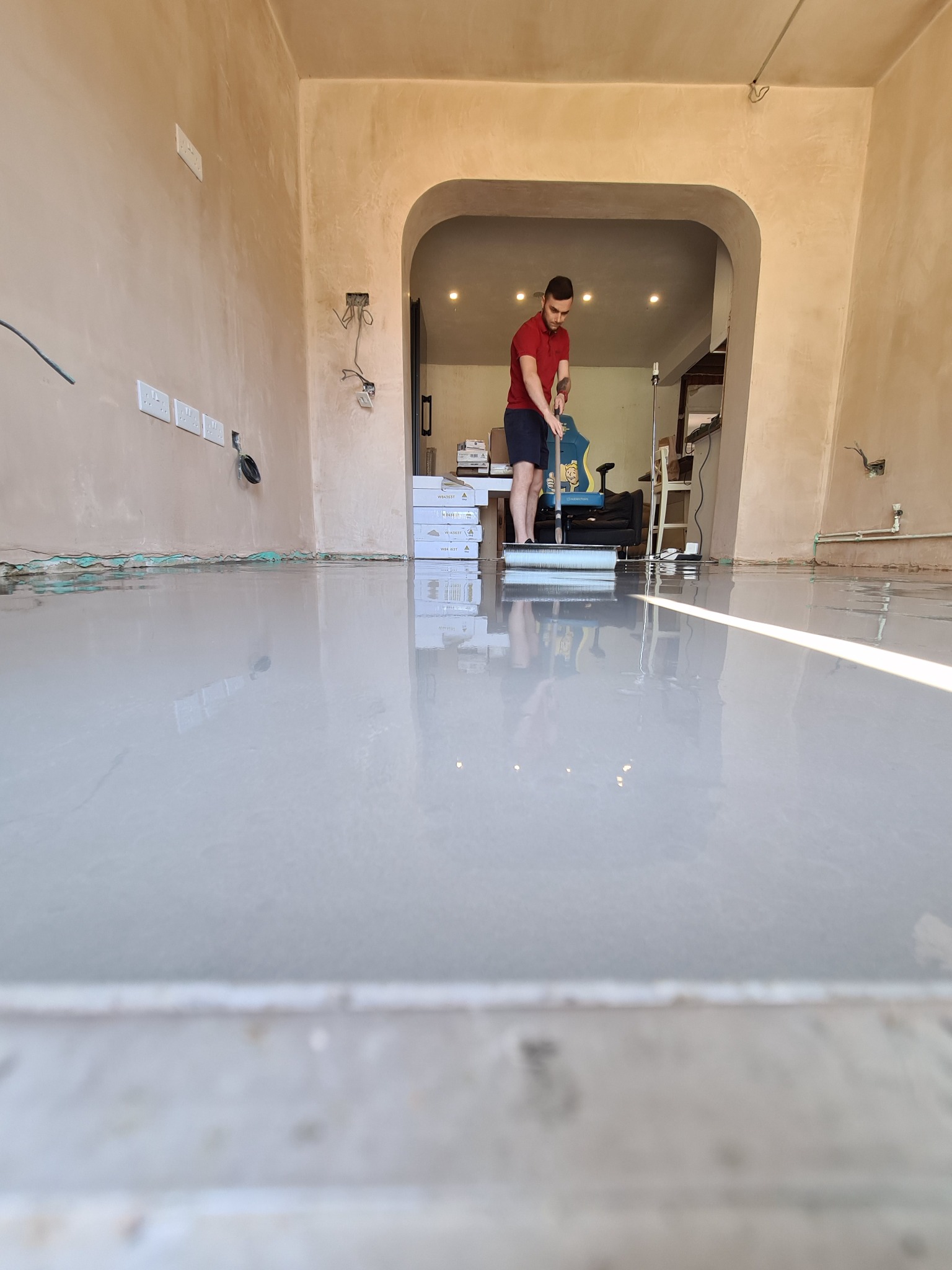 Mężczyzna rozprowadza szpachlą samopoziomującą masę na podłodze w pomieszczeniu z surowymi ścianami, widoczne puszki z masą i niezabezpieczone kable elektryczne.