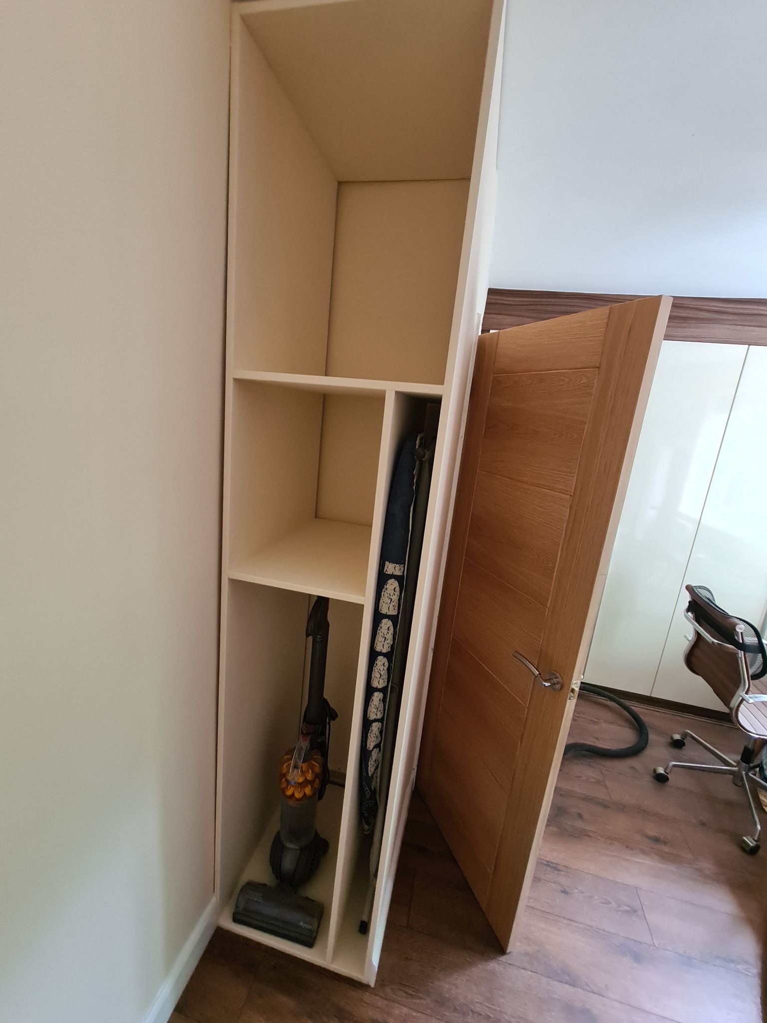 Wysoka, wąska, jasna szafa wnękowa z półkami, z odkurzaczem i deską do prasowania w środku, ustawiona obok drewnianych drzwi i biurowego krzesła na podłodze z paneli.