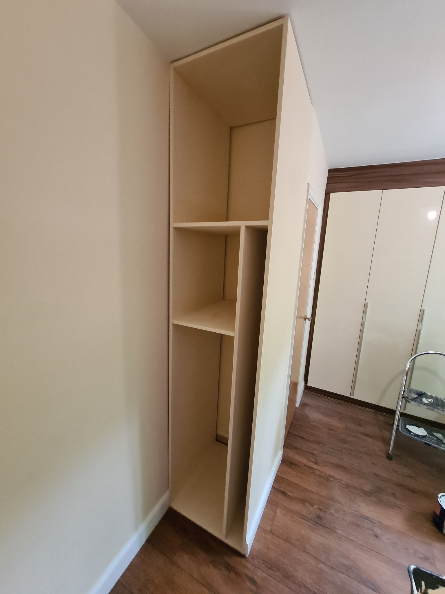 Wysoka, niedokończona, drewniana konstrukcja regału z półkami, ustawiona w rogu pokoju z panelami podłogowymi, obok białej szafy i drabiny malarskiej.