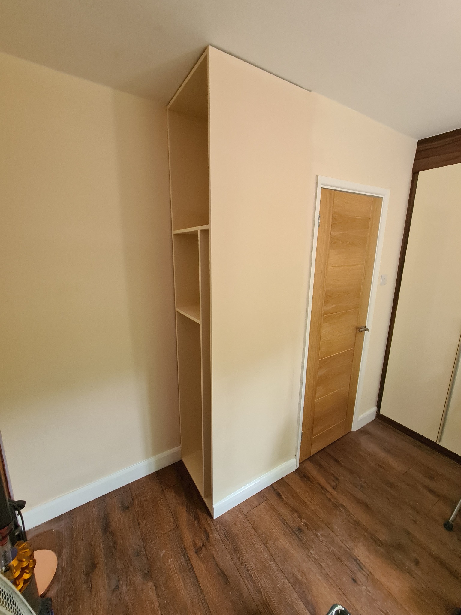 Wysoka, beżowa szafa wnękowa z półkami, zamontowana w rogu pokoju z drewnianą podłogą i drzwiami, widoczne listwy przypodłogowe.