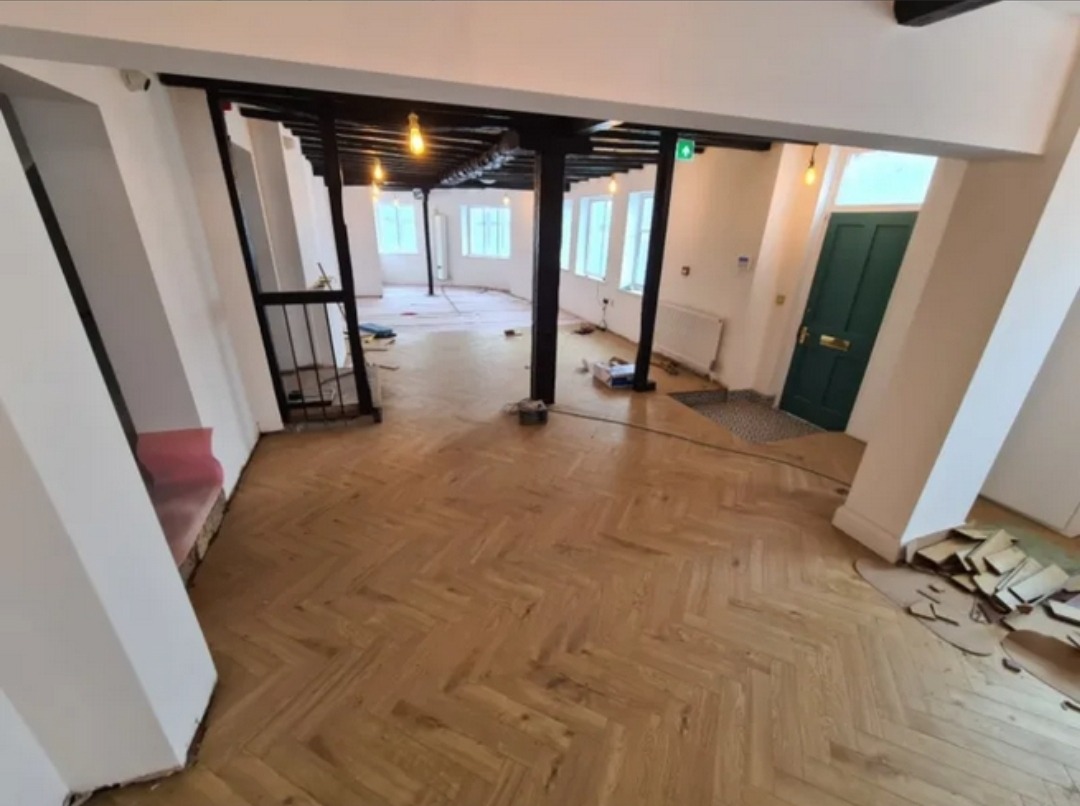 Jasna, nowa podłoga drewniana ułożona w jodełkę w przestronnym wnętrzu biurowym, z widocznymi fragmentami niezakończonego montażu i elementami wykończeniowymi.