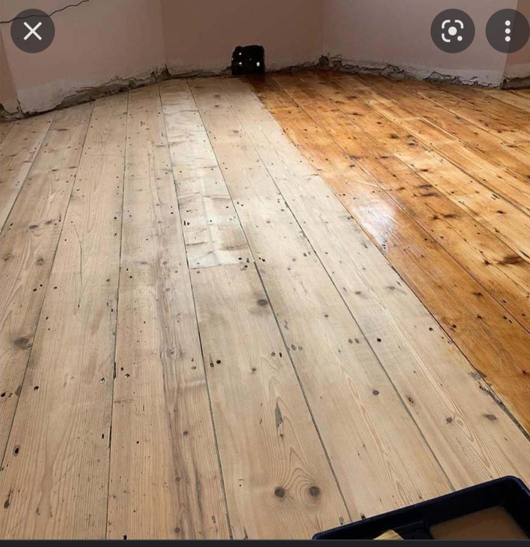 Drewniana podłoga w trakcie renowacji, widoczna różnica między surowym drewnem a polakierowaną powierzchnią, z widocznym czarnym pojemnikiem na lakier w rogu pomieszczenia.