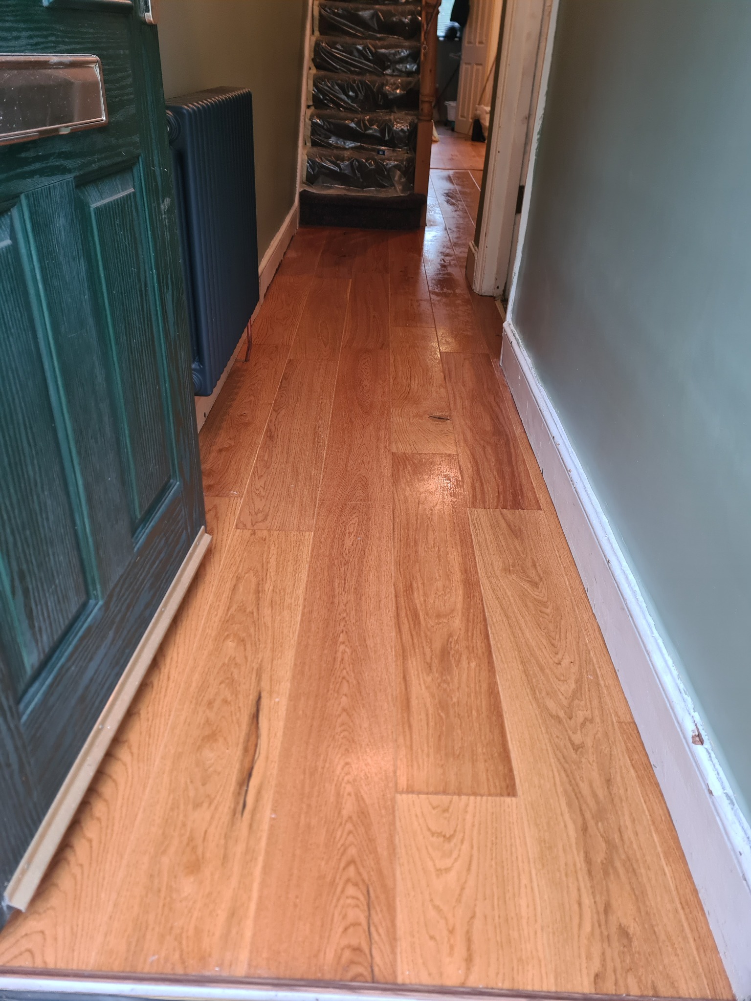 Świeżo ułożona, jasna podłoga drewniana w korytarzu, widoczna od progu wejścia, z fragmentem zielonych drzwi po lewej stronie i grzejnikiem w tle. Schody zabezpieczone folią ochronną.