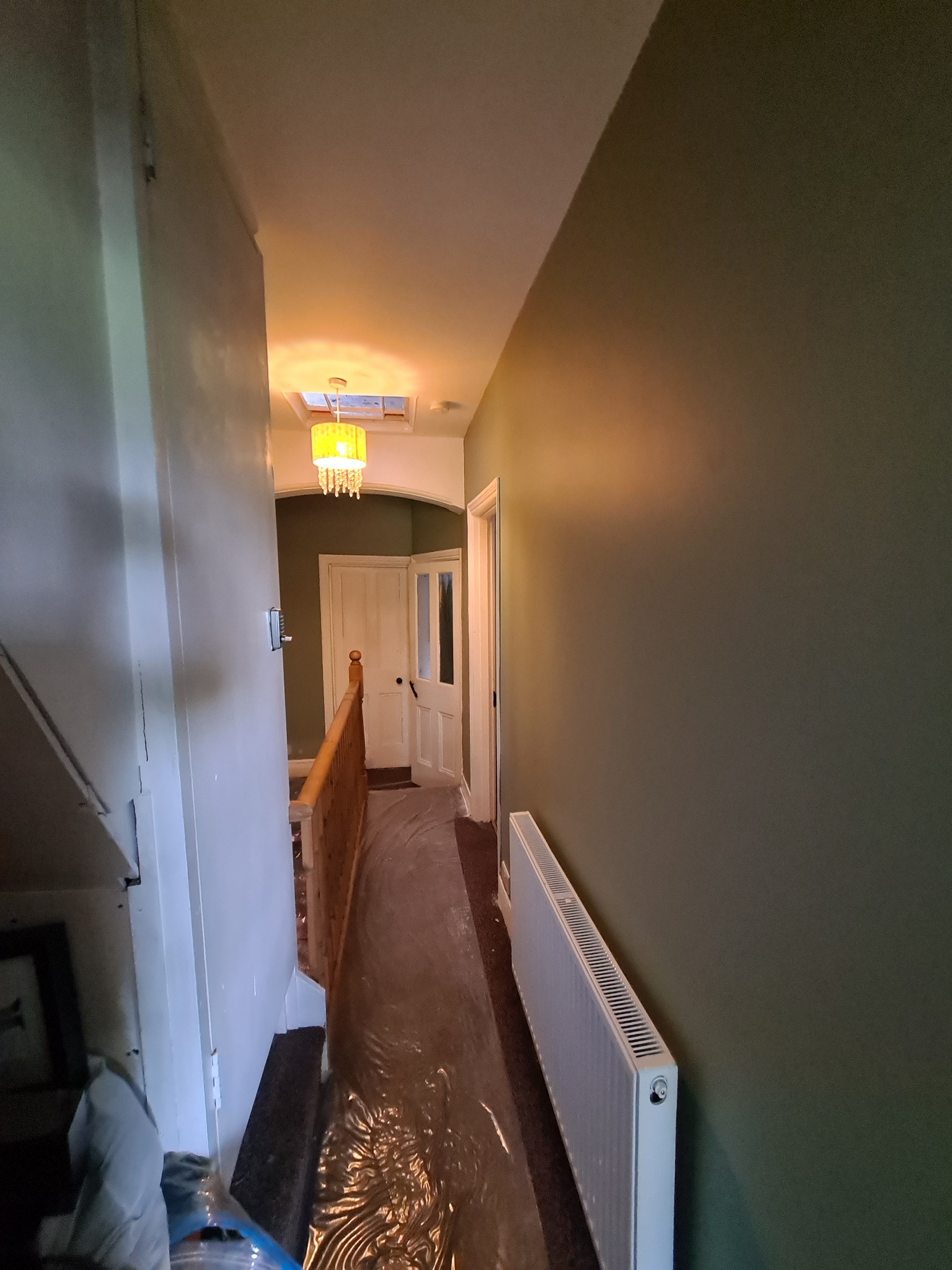 Korytarz po malowaniu ścian na oliwkowy kolor, podłoga zabezpieczona folią malarską, widoczny grzejnik i wisząca lampa z żółtym abażurem.