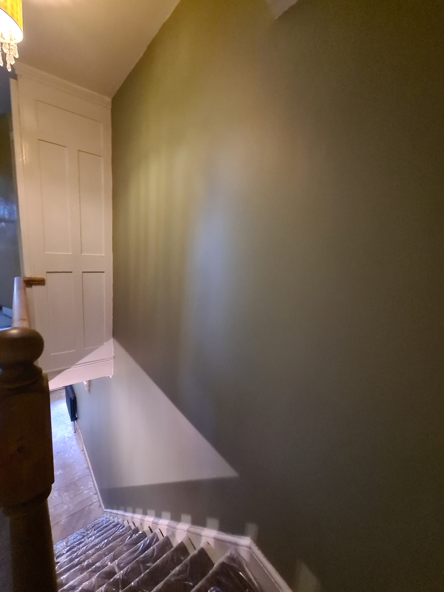 Świeżo pomalowana ściana w odcieniach zieleni, widoczna klatka schodowa zabezpieczona folią malarską, białe drzwi w tle, widok z góry.