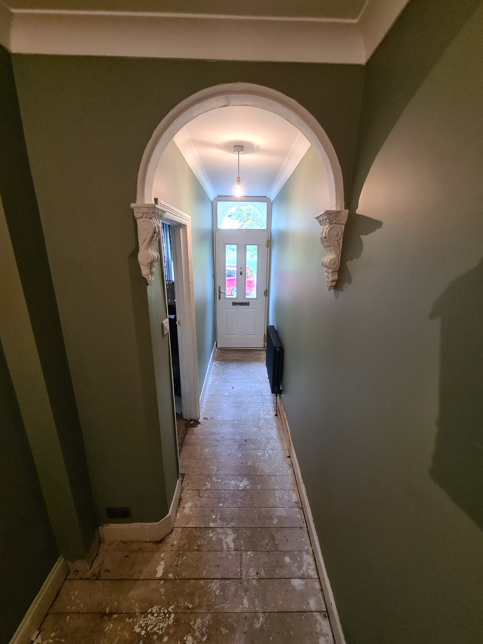 Korytarz w trakcie remontu: zielone ściany, surowa drewniana podłoga, biały łuk z dekoracyjnymi wspornikami prowadzący do białych drzwi wejściowych.