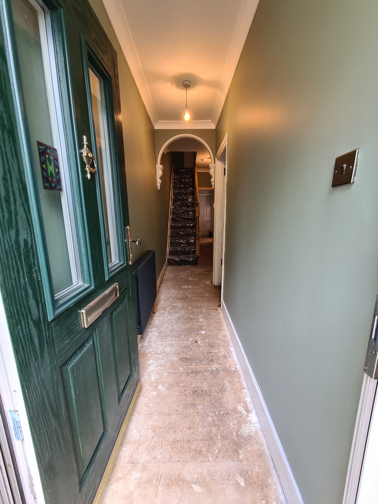 Korytarz w trakcie remontu: ściany świeżo pomalowane na oliwkowy kolor, zabezpieczone schody prowadzące na górę, stara podłoga przygotowana do położenia nowej, ciemnozielone drzwi wejściowe...