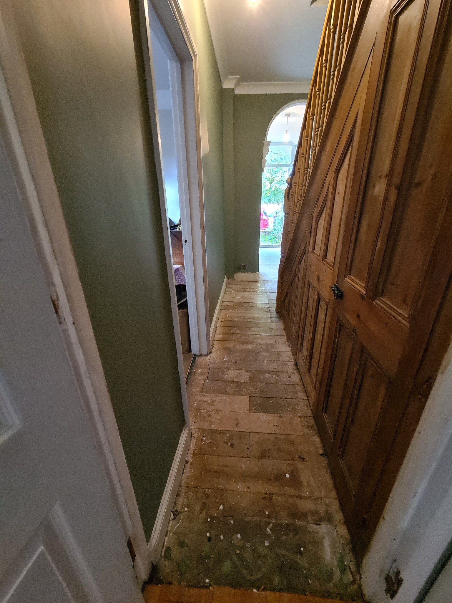 Korytarz w trakcie remontu: widoczne stare, częściowo usunięte deski podłogowe, ściany w odcieniach zieleni, drewniane schody po prawej stronie oraz drzwi po lewej stronie kadru.