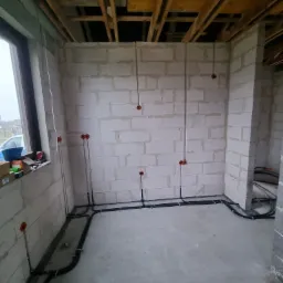 Surowy stan wnętrza z widoczną instalacją elektryczną: kable poprowadzone wzdłuż ścian z bloczków betonowych, puszki elektryczne zamontowane w ścianach, widoczne drewniane elementy konstrukcji stropu.