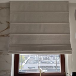 VTbud TETIANA LIUBACHIVSKA - Beżowa roleta rzymska w oknie kuchni, z widokiem na budynki. Złota bateria przy zlewie, marmurowe wykończenie ściany po lewej stronie. Stonowana kolorystyka.