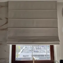 Beżowa roleta rzymska w oknie kuchni, z widokiem na budynki. Złota bateria przy zlewie, marmurowe wykończenie ściany po lewej stronie. Stonowana kolorystyka.