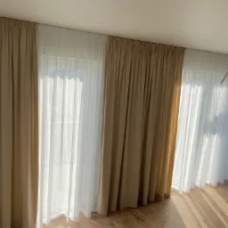 Beżowe i białe, eleganckie zasłony, upięte na karniszu, w jasnym pokoju z drewnianą podłogą. Delikatne światło słoneczne prześwieca przez firanki.