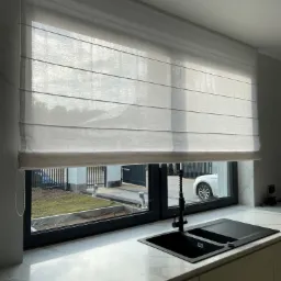 Jasna, płócienna roleta rzymska zamontowana w oknie nad czarnym zlewem kuchennym, widok na ogród z białym samochodem za oknem.