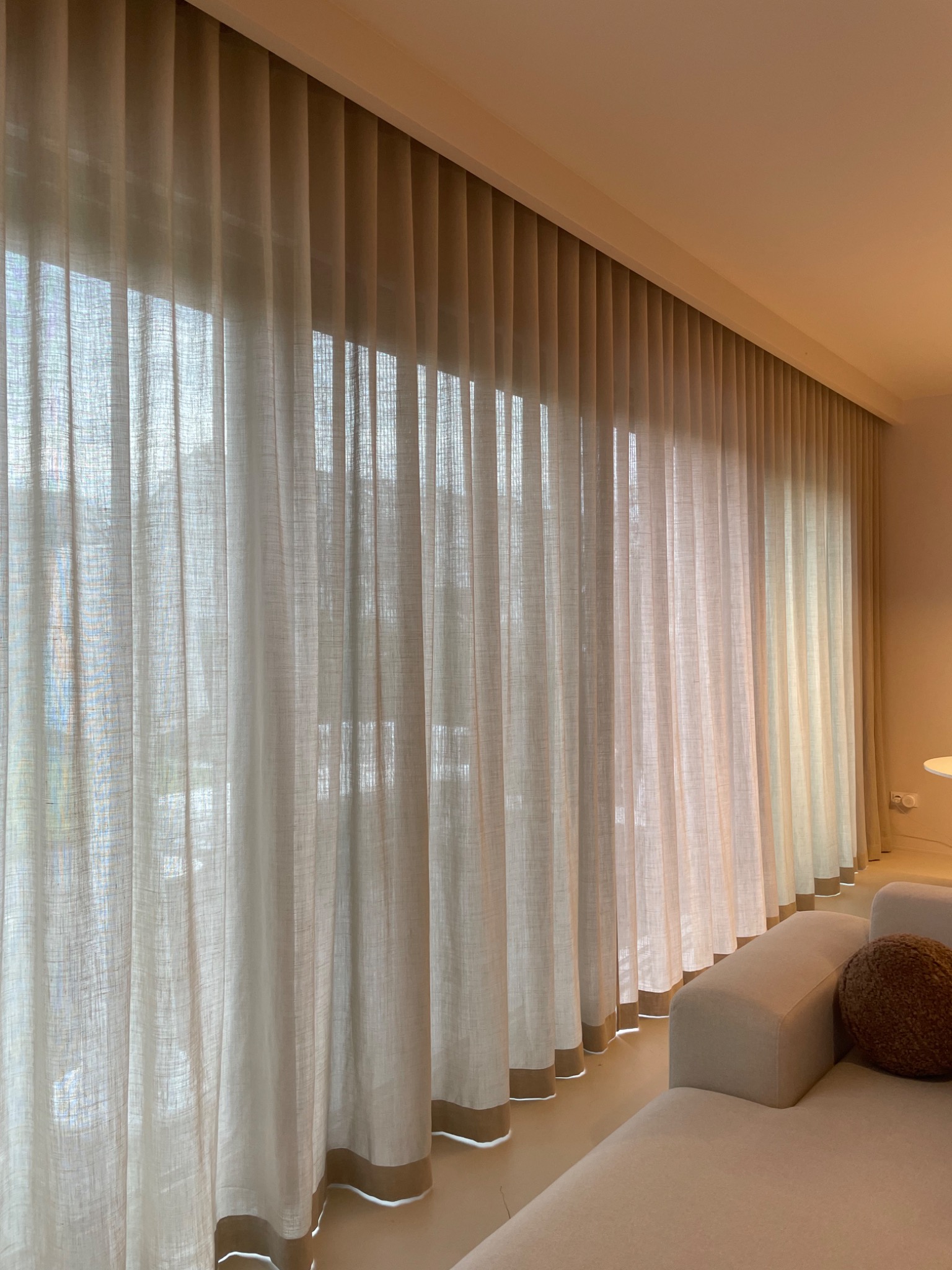 Długie, kremowe zasłony z delikatnym brązowym wykończeniem u dołu, wiszące w salonie z jasną podłogą i narożnikiem w tle.